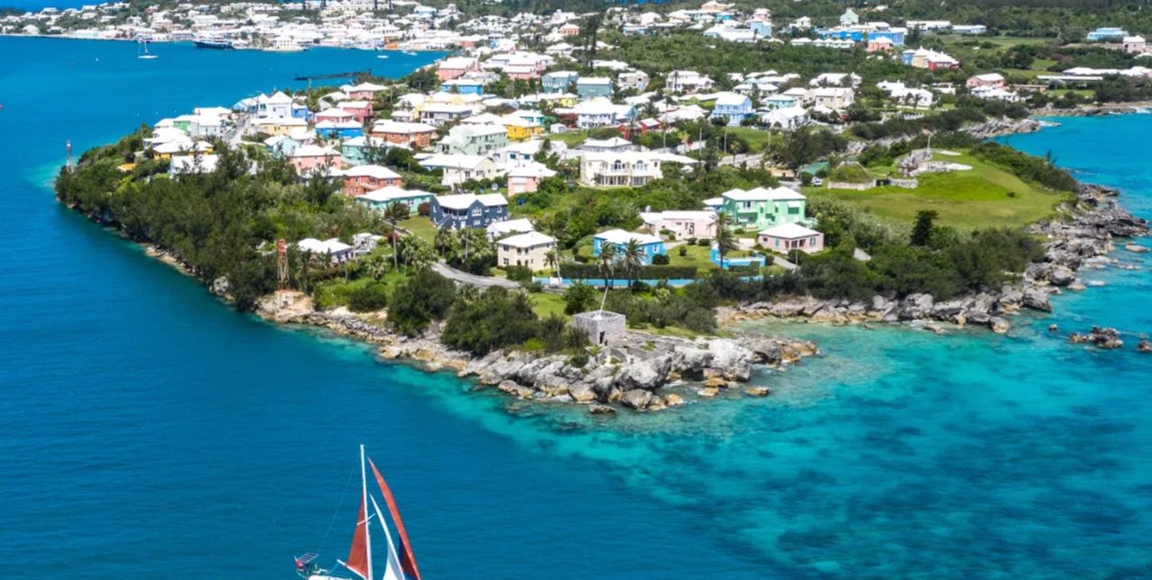 Buildings on island atop bright blue water, with blue and red sail boat in the foreground