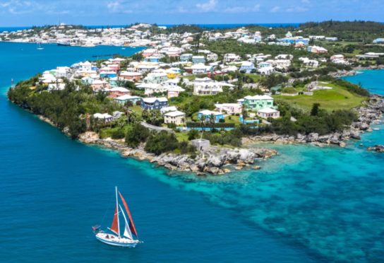 Buildings on island atop bright blue water, with blue and red sail boat in the foreground